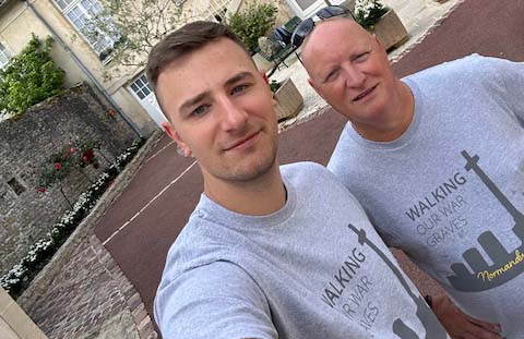 William and Stephen posing in grey CWGF Walking our War Graves t-shirts taking a selfie in front a small white fountain in a courtyard outside a Normandy hotel.