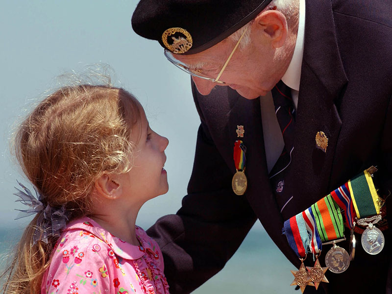 Elderly WW2 veteran in close conversation with a small girl in a pink dress. The veteran is wearing a military beret with cap badge, a white shirt and a blue blazer with a bar of military medals.