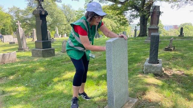 A women wearing a green hi-vis vest, grey cap, red shirt, and black trousers scrubs a CWGC headstone.