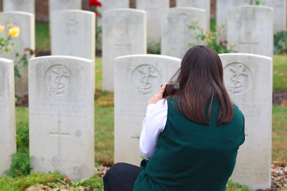 A young woman in a dark green vest and white shirt facing away from the camera takes pictures of CWGC headstones on her smartphone.