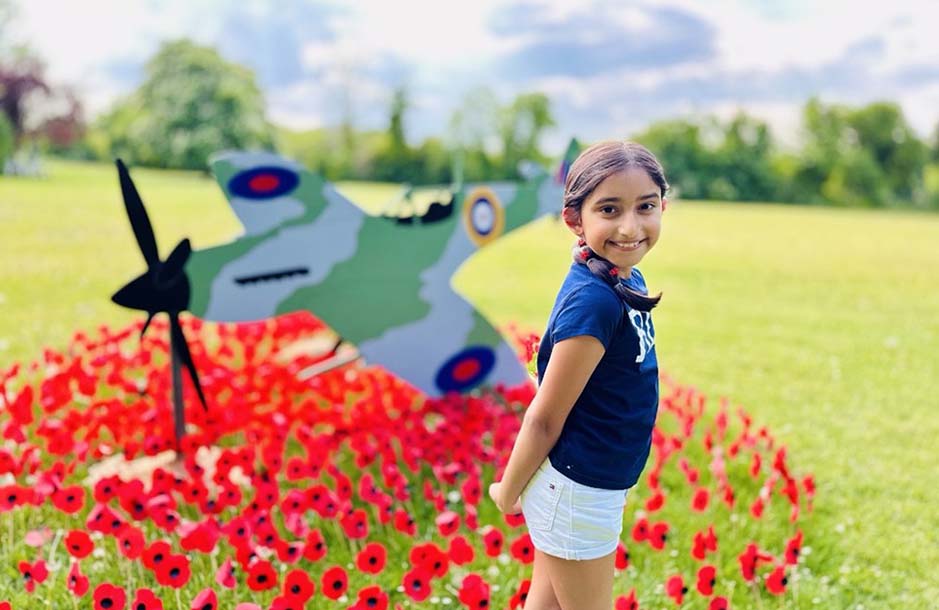 Small south Asian girl smiling in front of a wooden Spitfire sculpture sat over a bed of red poppies on a green school playing field.