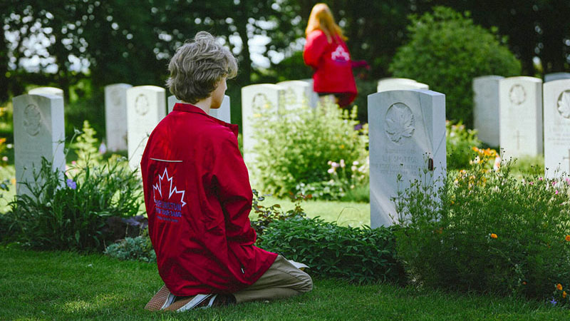 Youth in a red Canadian maple leaf jacket kneels contemplatively in front of a CWGC canadian war grave.