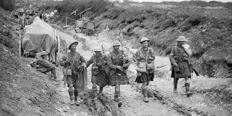 Four Black Watch soldiers walk down a muddy road during the battle of the Somme. Equipment and vehicles line the road.